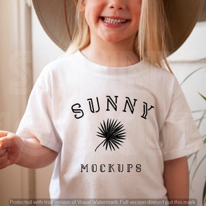 Op de afbeelding: Een wit T-shirt met een zwart grafisch ontwerp met de tekst "SUNNY" met daaronder een palmboomontwerp en de woorden "MOCKUPS" onder de palmboom.