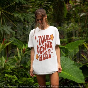 May include: A white t-shirt with a red and orange graphic that says "YOUR DESIGN HERE" with small yellow flowers around the text. The shirt is being worn by a person in a tropical setting.