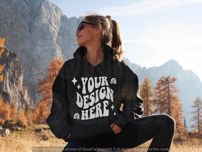 Pu&ograve; includere: Una felpa con cappuccio nera con il testo "YOUR DESIGN HERE" in bianco, con un arcobaleno e stelle. La felpa &egrave; indossata da una persona in piedi in un paesaggio montano.