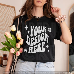 Puede incluir: Una camiseta negra con un gr&aacute;fico blanco que dice "YOUR DESIGN HERE" en una fuente retro. El gr&aacute;fico incluye estrellas e arco&iacute;ris.