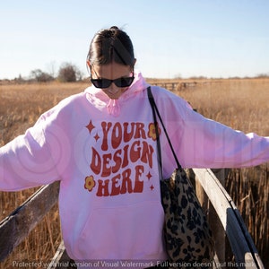 May include: A pink hoodie with the text "YOUR DESIGN HERE" in red and orange lettering. The hoodie has a flower design and stars on the text. The person is wearing sunglasses and is standing on a wooden walkway.