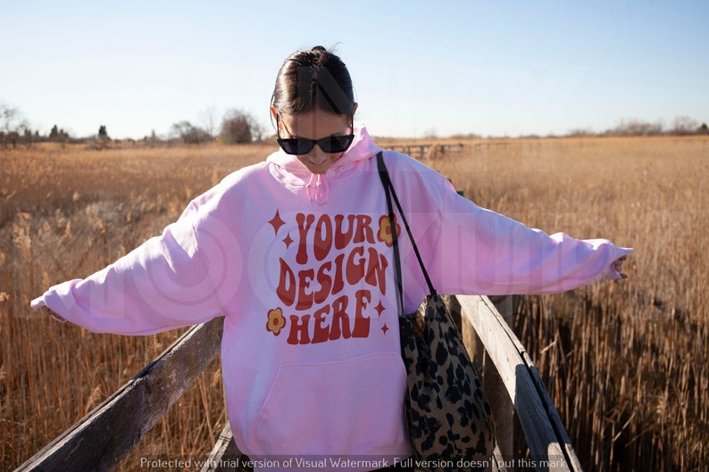 May include: A pink hoodie with the text "YOUR DESIGN HERE" in red and orange lettering. The hoodie has a flower design and stars on the text. The person is wearing sunglasses and is standing on a wooden walkway.