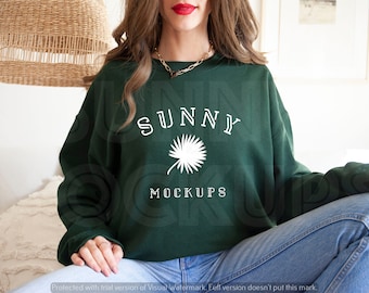 May include: A dark green sweatshirt featuring the text "SUNNY" and a palm tree graphic in white. The sweatshirt is being worn by a person sitting on a white surface.