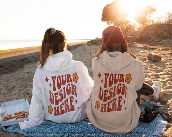 Puede incluir: Dos personas con sudaderas con capucha blancas y beige con el texto "YOUR DESIGN HERE" en rojo y amarillo en la espalda. Están sentadas en una playa con una caja de pizza delante de ellas.