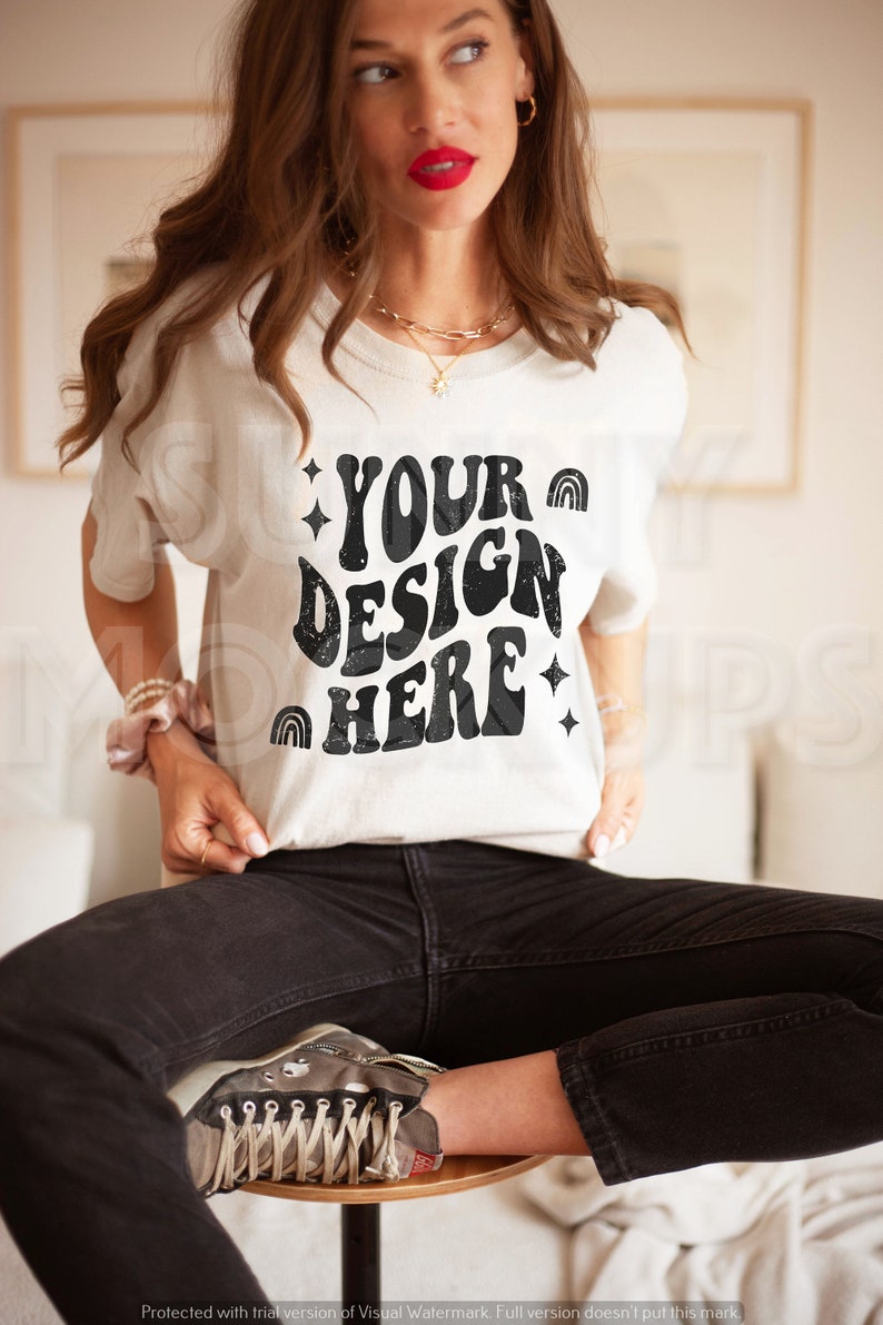 May include: A white t-shirt with a black graphic that says "YOUR DESIGN HERE" in a retro font. The graphic includes stars and rainbows. The shirt is being worn by a person with long brown hair and red lipstick. The person is sitting on a stool with their legs crossed.