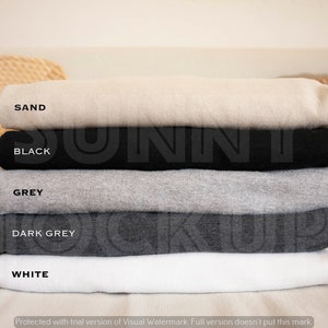 May include: A stack of five folded t-shirts in different colors: sand, black, grey, dark grey, and white. The shirts are folded neatly and stacked on top of each other.
