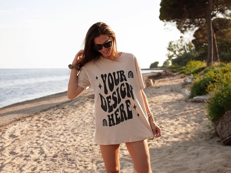 Puede incluir: Una mujer lleva una camiseta beige con el texto "YOUR DESIGN HERE" impreso en negro. La camiseta tiene un estilo de fuente retro y groovy. Est&aacute; de pie en una playa de arena con un oc&eacute;ano azul de fondo.