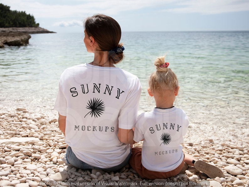 May include: Two people wearing white t-shirts with the text "SUNNY MOCKUPS" and a palm tree graphic on the back. The people are sitting on a beach with their backs to the camera, looking out at the ocean.