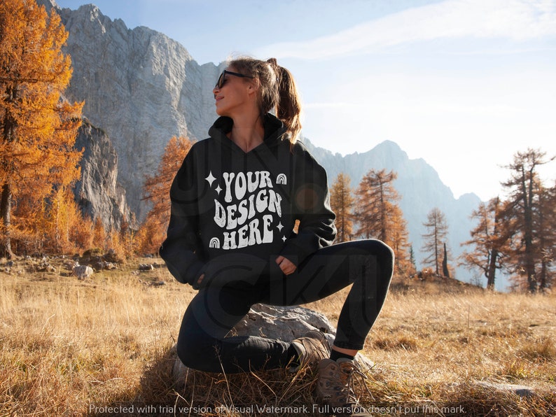 Pu&ograve; includere: Una felpa con cappuccio nera con una grafica bianca che dice "YOUR DESIGN HERE" in un font retr&ograve;. La felpa &egrave; indossata da una persona seduta su una roccia in un paesaggio montano.