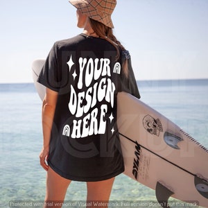 May include: A black t-shirt with the text "YOUR DESIGN HERE" in white, featuring a rainbow and stars. The shirt is being worn by a person standing on a beach with a surfboard.