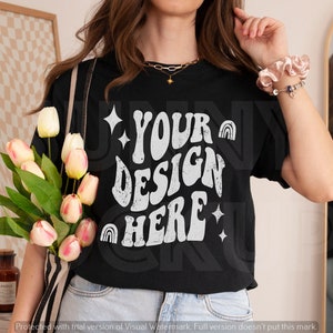 Puede incluir: Una camiseta negra con un gr&aacute;fico blanco que dice "YOUR DESIGN HERE" en una fuente retro. El gr&aacute;fico tambi&eacute;n incluye estrellas e arco&iacute;ris.