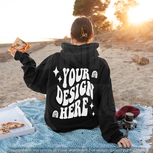 May include: A black hoodie with a white design on the back that says "YOUR DESIGN HERE" with stars and rainbows. The hoodie is being worn by a person sitting on a blanket on a beach.