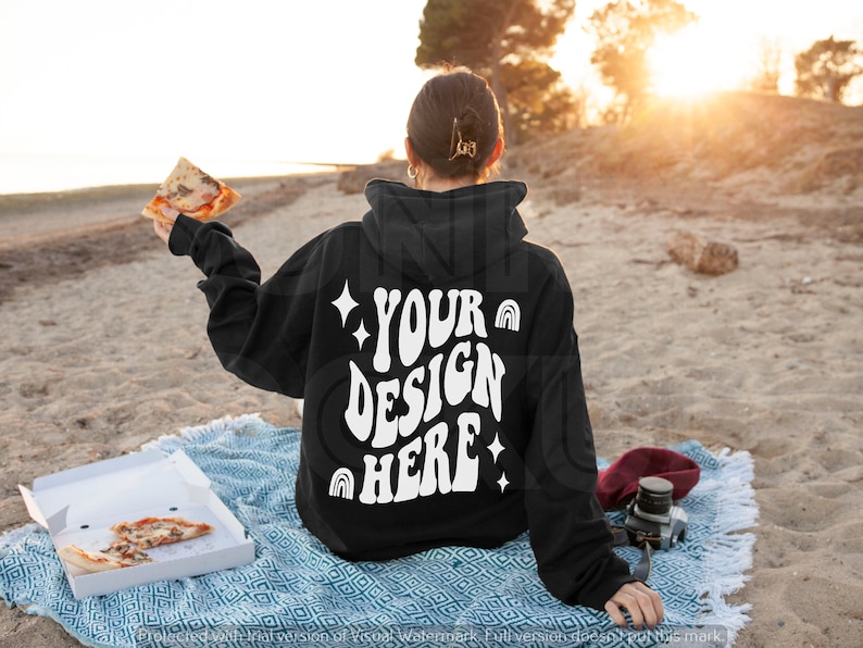 May include: A black hoodie with a white design on the back that says "YOUR DESIGN HERE" with stars and rainbows. The hoodie is being worn by a person sitting on a blanket on a beach.