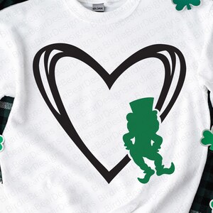 May include: A white long-sleeve sweatshirt featuring a large black outlined heart graphic. Inside the heart, a solid green leprechaun silhouette wearing a top hat is positioned on the right side. Three green shamrock icons are placed around the heart.