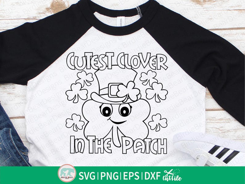 Pu&ograve; includere: Una maglietta bianca e nera in stile raglan con un design per la festa di San Patrizio. La grafica include un trifoglio sorridente con un cappello da folletto, circondato da trifogli, e la scritta "Cutest Clover in the Patch."