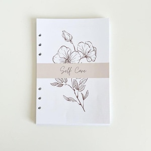 May include: A white notepad with a floral illustration and the words "Self Care" printed on a light brown band. The notepad has three holes on the left side for binding.