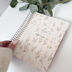 May include: A spiral-bound budget planner with a floral design in soft beige, pink, and green tones. The words "Budget Planner" are printed on the cover. The planner is open, revealing the interior pages.