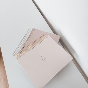 May include: Two light pink spiral-bound notebooks with gold-colored wire binding. The top notebook has the year "2026" printed on the cover in a simple, handwritten font. The notebooks are stacked on a light gray surface.