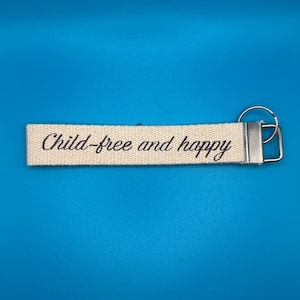 May include: A beige fabric keychain with the words "Child-free and happy" in black script. The keychain has a silver metal clasp and a silver key ring. The background is a solid blue.