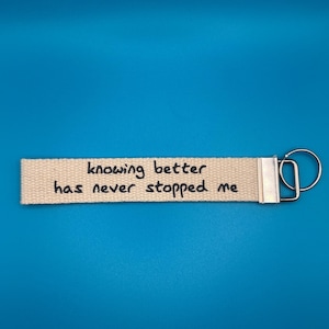 May include: A beige fabric keychain with the text "knowing better has never stopped me" in black lettering. The keychain has a silver-coloured metal clasp and ring, set against a bright blue background.