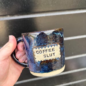 May include: A handmade ceramic mug with a glossy, mottled blue and dark brown glaze. The mug features a rectangular, cream-coloured textured plaque with scalloped edges that has the words COFFEE SLUT stamped in black capital letters on the front.