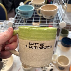 May include: A ceramic mug with a light green glazed rim and a textured beige body. The front of the mug features the black text DONT BE A CUNTASAURUS. The mug has a handle on the left side and a smooth interior finish, showcasing a handmade aesthetic.