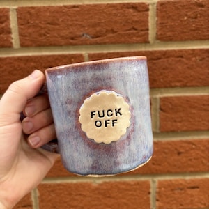 May include: A ceramic mug with a blue and purple glaze. The mug features a cream-colored, scalloped-edge emblem with the words "FUCK OFF" in black, centered on the front. The mug is held by a hand against a brick wall.