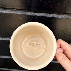 May include: A cream-coloured ceramic mug with a smooth, glazed interior. The base of the mug features the word TWAT embossed in capital letters. The mug has a simple, cylindrical shape and a glossy finish that reflects light from the surface.