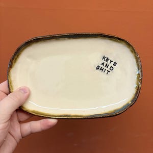 Op de afbeelding: Een ovaal keramieken dienblad met een crèmekleurig midden en een donkere, rustieke bruine rand. Het oppervlak bevat de zwarte gestempelde tekst "KEYS AND SHIT" aan de rechterkant. Het dienblad heeft een onregelmatige, handgevormde rand.