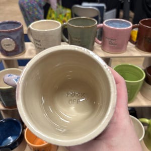 Op de afbeelding: Een verzameling handgemaakte keramische mokken in verschillende kleuren, waaronder blauw, groen, roze en beige. De binnenkant van een mok toont de zin "FUCK OFF". Andere mokken hebben tekst. De mokken worden op een houten plank tentoongesteld.