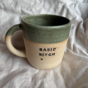 May include: A handmade ceramic mug with a green upper section and a beige lower section. The words "BASIC BITCH" are printed in black on the beige section. The mug has a curved handle.