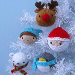 Op de afbeelding: Vijf gebreide kerstversieringen hangen aan een witte kunstkerstboom. De versieringen zijn een rendier, een pinguin, een sneeuwpop, een kerstman en een elf. De versieringen zijn allemaal wit, rood, blauw en bruin.