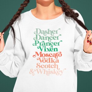 Könnte beinhalten: Weißer Pullover mit einem grünen und rosafarbenen Grafikdruck mit der Aufschrift "Dasher Dancer Prancer Vixen Moscato Vodka Scotch & Whiskey".