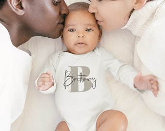 May include: A baby wearing a white onesie with the name "Brittany" and a large grey letter "B" on the front. The baby is being kissed on the cheeks by a man and a woman.