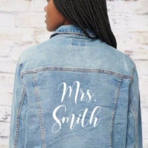 Pode incluir: Uma jaqueta de ganga azul claro com o texto "Mrs. Smith" em branco nas costas.