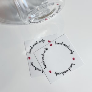 May include: Four white square stickers with the text "hand wash only" written in black around the edge of each sticker.  Each sticker has four small red hearts around the edge.
