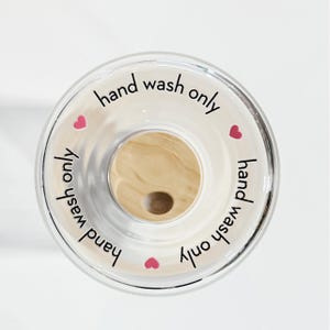 May include: A clear glass bottle stopper with a wooden top and the text "hand wash only" written around the edge. There are four pink hearts around the edge of the stopper.