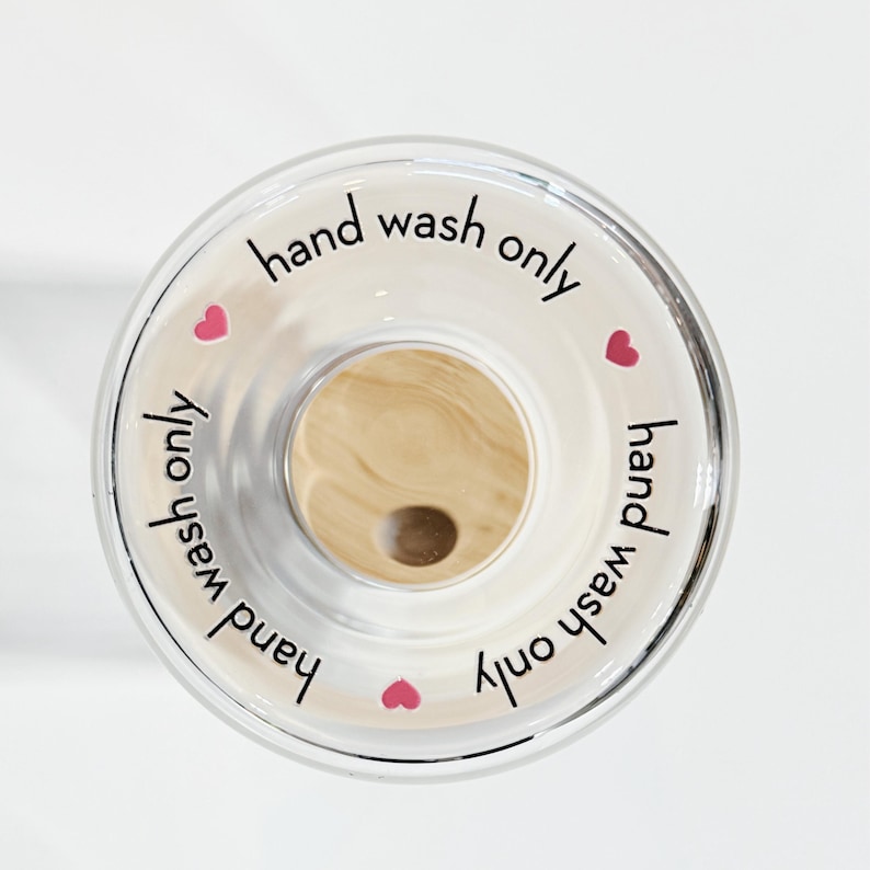 May include: A clear glass bottle stopper with a wooden top and the text "hand wash only" written around the edge. There are four pink hearts around the edge of the stopper.