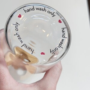 May include: A clear glass cup with a brown wooden handle. The cup has the text "hand wash only" written around the rim with a small red heart on each side.