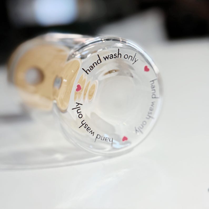 May include: Close-up of a clear glass bottle with a hand wash only label. The label has a pink heart on each side and the text "hand wash only" is repeated twice.