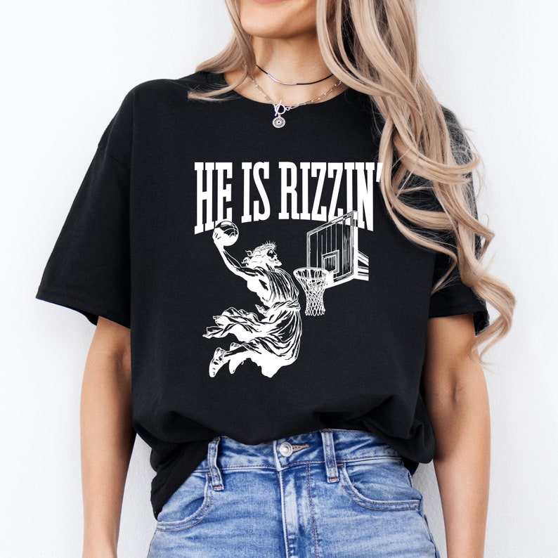 May include: Black t-shirt with a white graphic of a person dunking a basketball with the text "HE IS RIZZIN'" above the graphic.