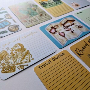 May include: A selection of travel-themed cards with diverse designs. Some cards display maps, globes, and travel illustrations in gold and brown hues. Other cards have lined spaces for writing, with phrases such as "Travel Diaries" and "your next adventure".