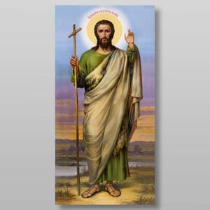 Puede incluir: Una pintura de icono religioso de Juan el Bautista, representado con una larga barba, vistiendo una túnica verde y una capa marrón, sosteniendo un bastón con una cruz en la parte superior. Está de pie en un paisaje desértico con un río en el fondo. La pintura tiene un halo alrededor de su cabeza y el texto "Святый пророкъ и Предтеча Иоаннъ" está escrito en la parte superior.