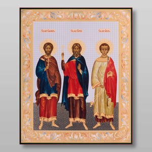 May include: An icon depicting three saints, each with a halo and holding a cross. The saints are standing in a row, with the saint on the left wearing a blue robe and red sash, the saint in the middle wearing a red robe and blue sash, and the saint on the right wearing a yellow robe and red sash. The icon is framed in gold and has a floral design.