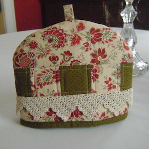 May include: A cream-colored tea cozy with a floral pattern in shades of red, green, and brown. The cozy has a lace trim and a green quilted section.
