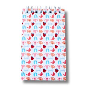 May include: A spiral-bound notepad with a repeating pattern of cartoon human organs, including hearts, lungs, stomachs, and reproductive systems. The organs are in various colours, such as red, pink, and blue, set against a light blue background. The notepad has a silver spiral binding.