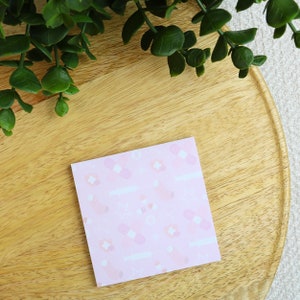 Könnte beinhalten: Ein rosa Haftnotizblock mit einem sich wiederholenden Muster aus weißen Verbänden, Blumen und Spritzen. Der Haftnotizblock liegt auf einer hellbraunen Holzoberfläche.