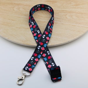 Peut inclure: Lanière noire avec un motif blanc et rouge de cœurs, stéthoscopes et bonnets d'infirmière. La lanière a un clip argenté à une extrémité et un clip en plastique noir à l'autre extrémité.