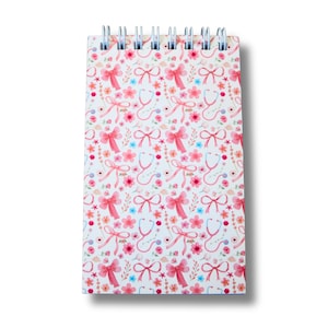 May include: A spiral-bound notepad with a white cover adorned with a repeating pattern of pink bows, stethoscopes, and floral elements. The design incorporates shades of pink, blue, and orange. This notepad is suitable for jotting down notes and has a feminine style.