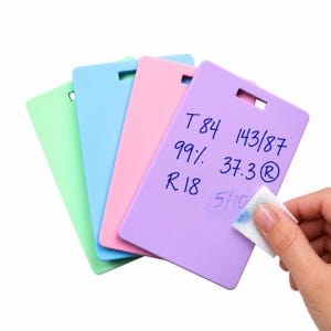 May include: Four rectangular plastic boards in light green, blue, pink, and purple, each with a slot at the top. The purple board features handwritten blue ink text: T 84 143/87 99% 37.3 R 18 5/10. A white cloth is wiping away some of the blue ink.